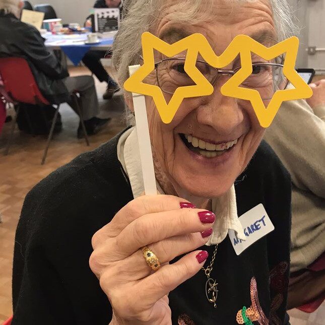 Age UK Enfield charity photo of elderly woman smiling while holding yellow star glasses to her face