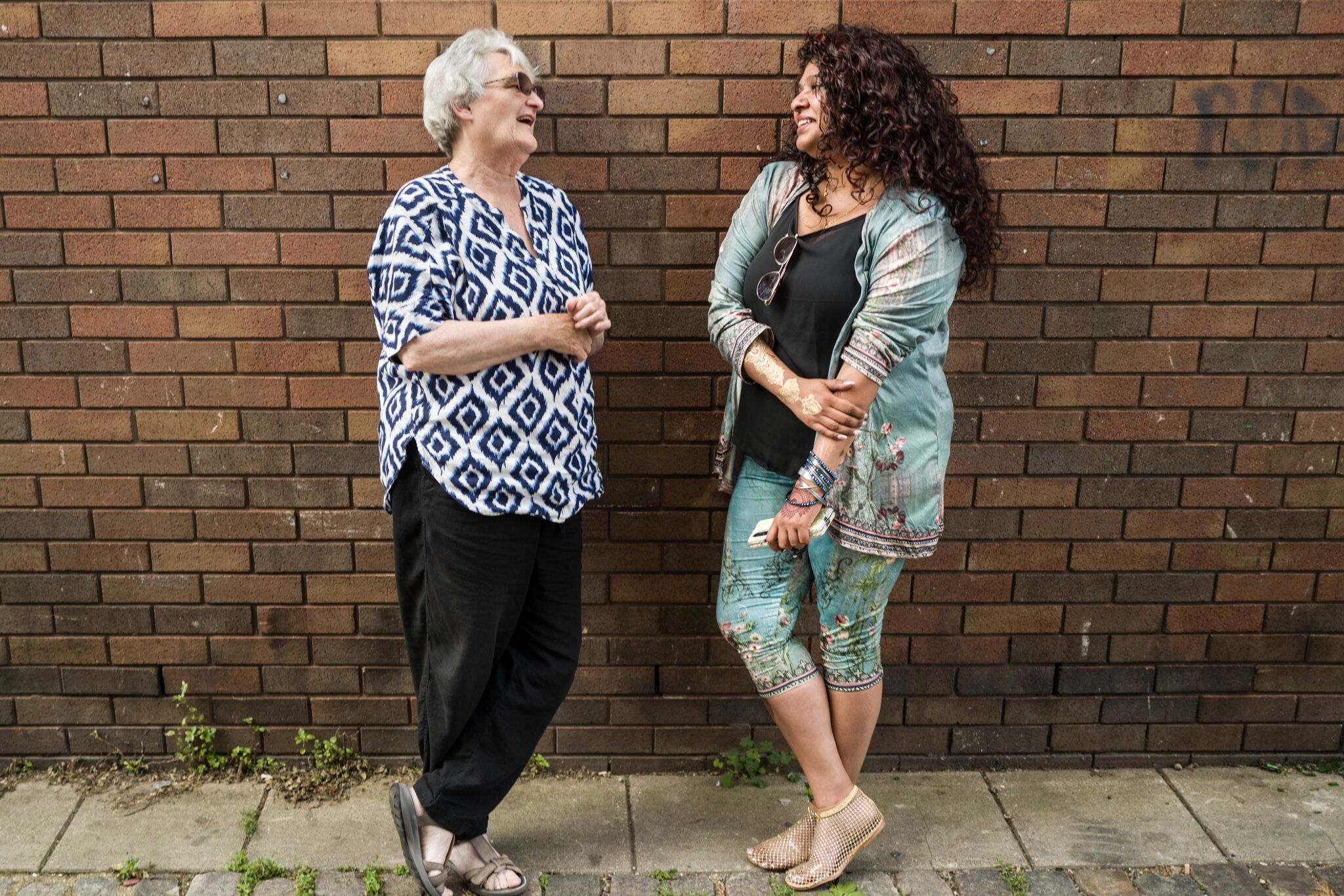 two women smiling and talking whilst leaning against a brick wall