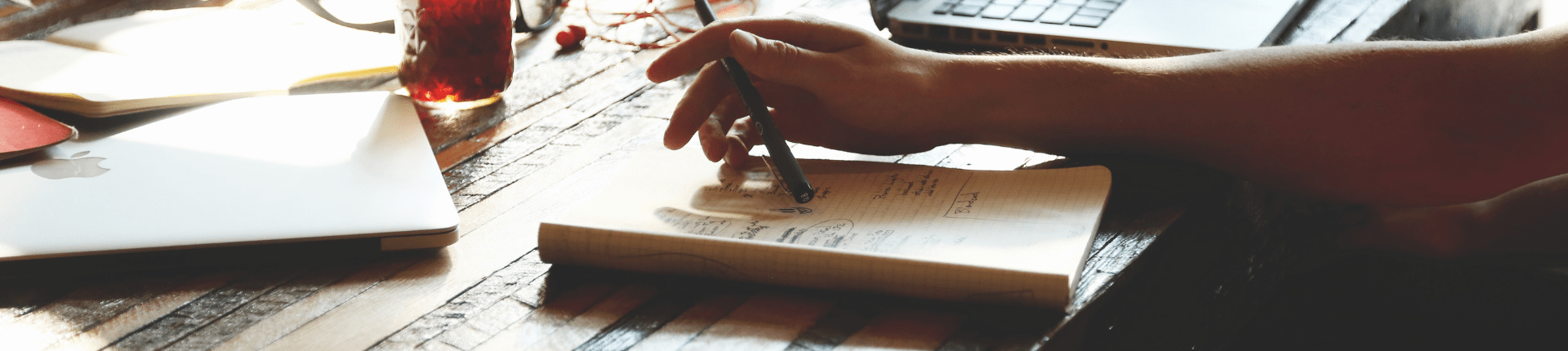 Arm of a person holding a pen with a note book and a laptop computer. Good Merger Guide 5 of 7 Feasibility study website banner