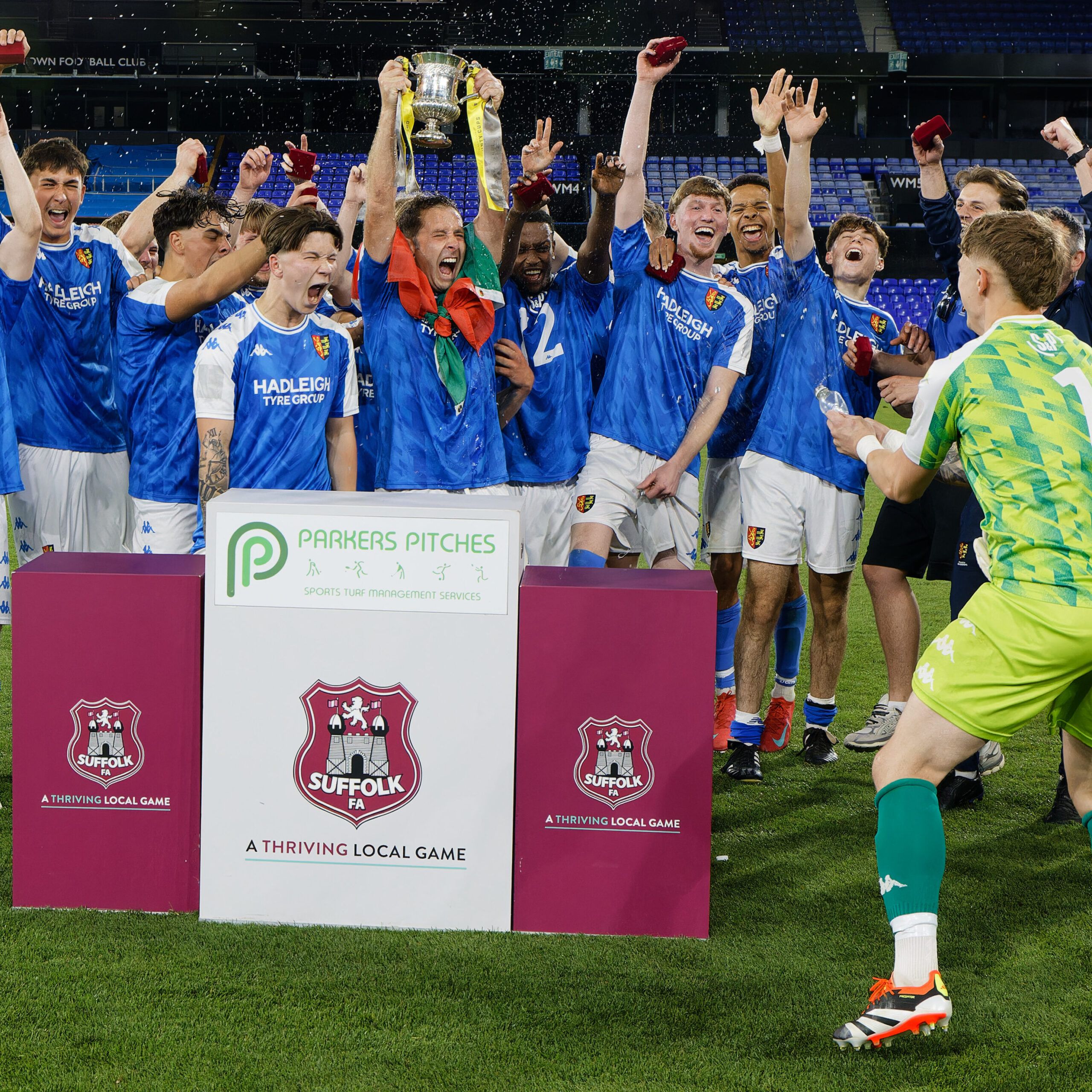 Suffolk FA Junior Cup male players wearing blue and white kit on the pitch, cheering and holding cup in the air.
