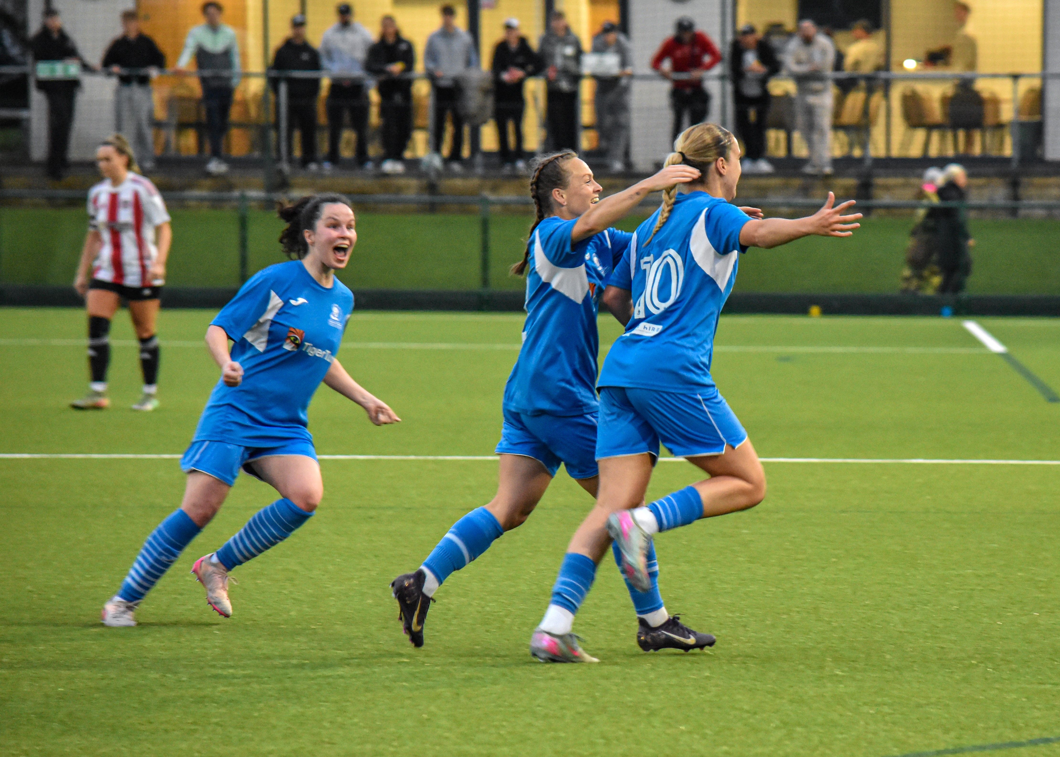 3 girls in football kits on a football field, one girl is smiling and one girl has her arm around the other celebrating