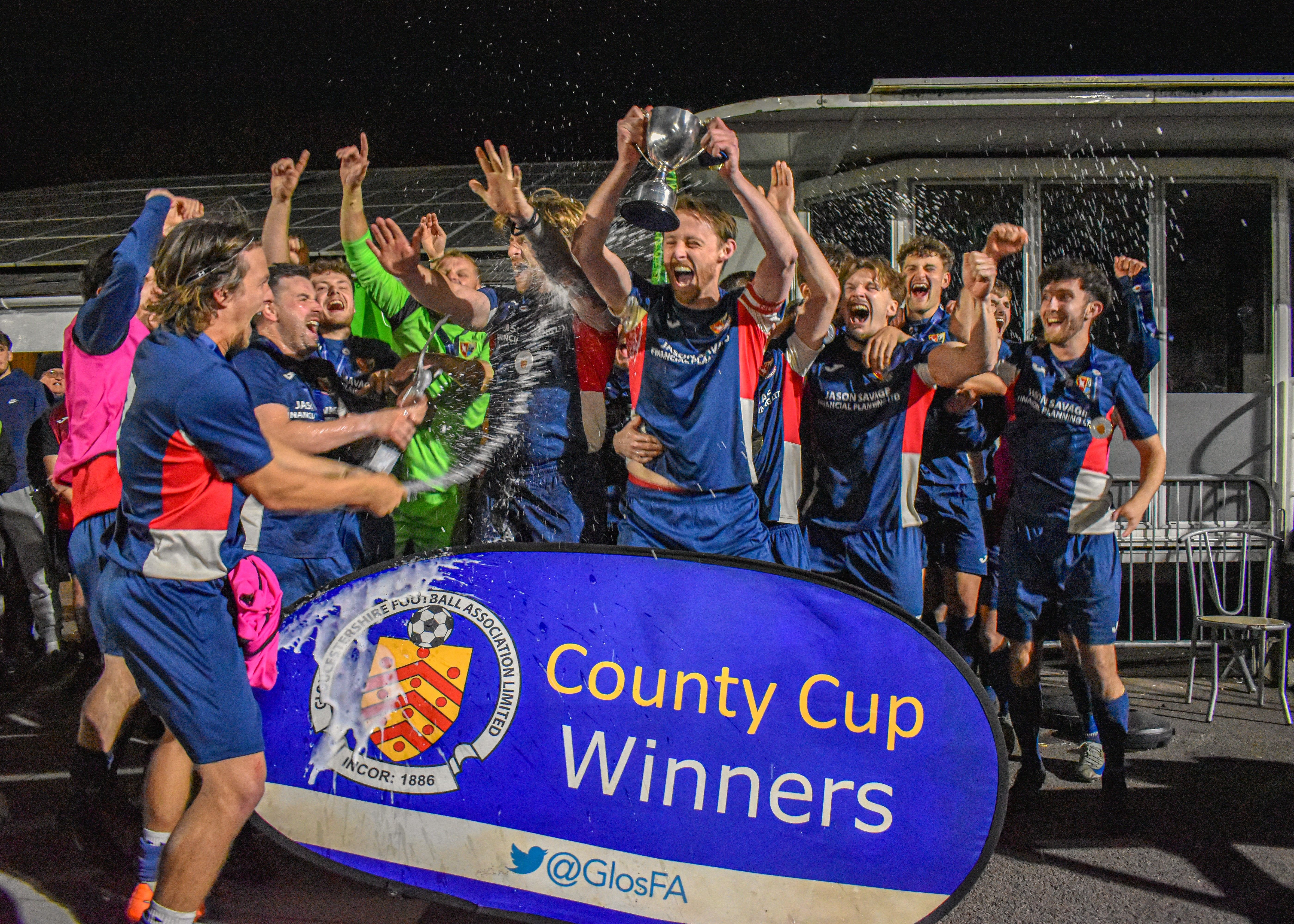 A group of men in football kits celebrating with a trophy and standing behind a banner that says County Cup. Winners with Gloucestershire FA logo