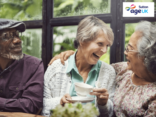 3 elderly people drinking tea and smiling in front of a large window. Age UK Sutton